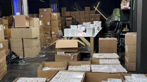 Cardboard boxes containing empty and pre-filled weight-loss jabs are stacked in a warehouse with strip lighting.
