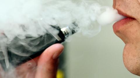 A black vape is held in the hand of a man who is exhaling smoke. 