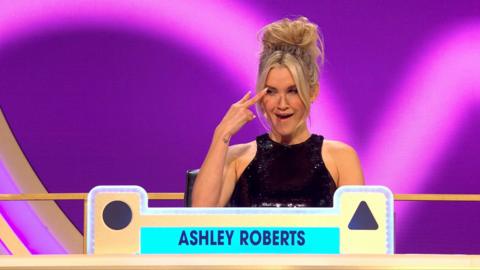 Ashley Roberts in the Blankety Blank studio - she holds two fingers to her eyes, and wears a black sleeveless top