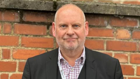 Jonathan Nunn with short hair and a beard, standing in front of a brick wall