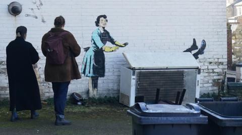 Two people looking at the artwork which shows a spray-painted woman pushing someone into a freezer.