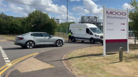 A sign including the words Moog Wolverhampton is on grass. A person is standing by a white van, which is on a road near a silver car.