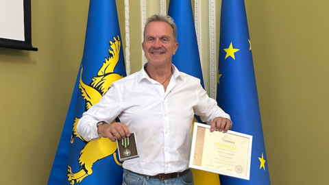 Prof Mark Hannaford standing in front of three flags holding a medal and a framed award document