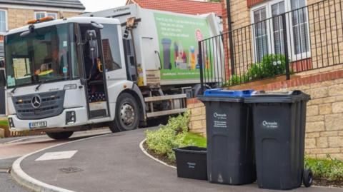 A recycling lorry pictured next to two black bins and a black recycling box