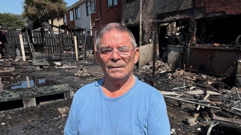 Davy Haighton, a man with short grey hair and silver-framed glasses stands in front of the wreckage of two terraced houses which have been gutted by fire. He is wearing a light blue crew-neck t-shirt