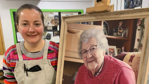 Megan Hunter wearing an apron, standing beside an easel with the painting she has created of her Nanna. It looks like a photograph because it is a realism painting. In the portrait, her Nanna has grey hair and is wearing a pink jumper and glasses.