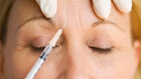 A close-up of a needle put on woman's forehead and the top of the nose, with the fingertips of a gloved hands just showing above the injection point.