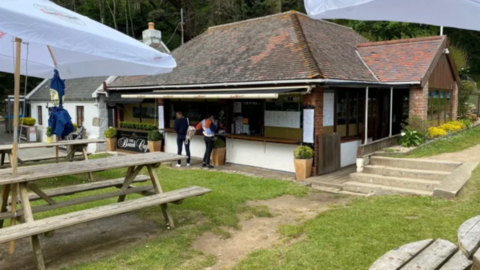A one-storey building with two people queuing outside to order food from the cafe. It is surrounded by green grass and small potted green bushes. There are picnic benches and large umbrellas surrounding the area. 