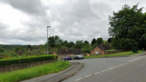 A road junction, with cars parked on the street which is dipping down a hill. There are street lights and houses as well as a pavement and a large tree near the junction.