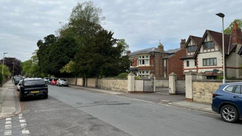 A wide residential street with large houses with gates in front of them on the left. A number of cars are parked along the road to the left of the road. Large green trees form a barrier in front of large houses further down the road.