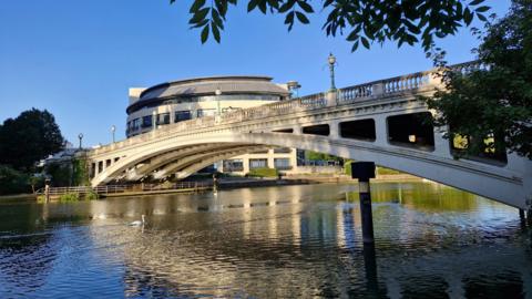 A sunny day with a bridge dominating the image going over a waterway. There is a round-shaped building in the background and overhanging trees in the foreground.