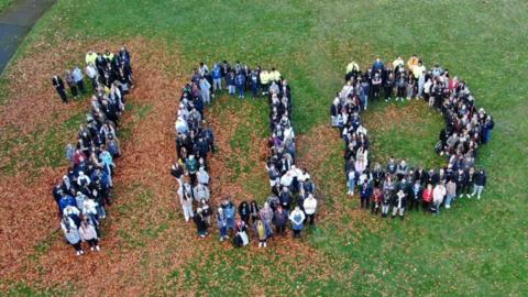 An aerial view of people gathered together to display the number 100. They are standing on grass, which is partially covered in brown leaves.