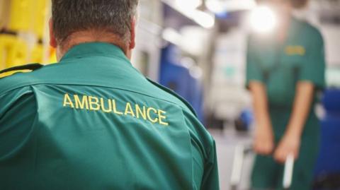 Two ambulance workers - one seen from behind and one blurred in the background. They are wearing green uniforms, with yellow lettering. The man in the foreground has 'Ambulance' written on his back.