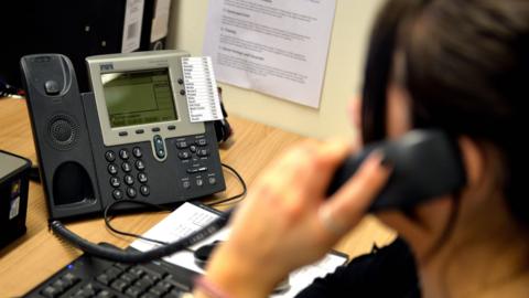 A woman sitting at a desk with a black computer and keyboard. She is speaking on a black telephone with the phone held to her left ear.