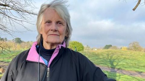 Maggie Chapman is standing outside and the golf course can been to one side of her. She is wearing a dark jacket with a pink collar and a blue jumper underneath.