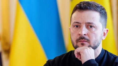 Zelensky, sitting in front of a Ukrainian flag, rests his chin on his hand, looking deep in thought