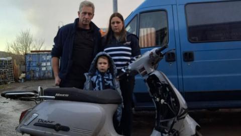 Becca, John and Joseph are standing behind the Neco Dinno scooter which was stolen last week. It has been damaged and the family look unhappy. Behind them is a blue van.