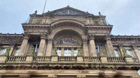 An external shot of the upper floor Birmingham Council House, which is a sandstone building with moulded columns and a balcony. Statues of lions line the balcony on plinths and a central arch sits behind it, featuring a mosaic. A pale blue sky can be seen above.