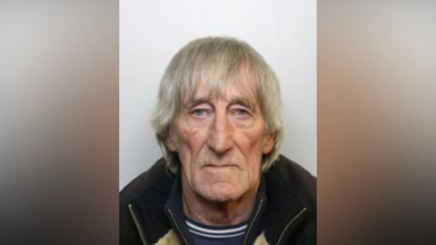 A mug shot of a man with long grey hair, blue eyes, wearing a navy jumper with white stripes and a jacket.