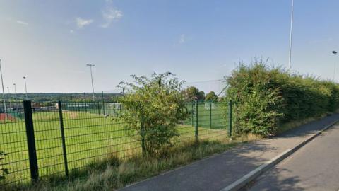The playing fields of Weydon School, viewed over the fence from the public road, with floodlights standing in the distance.