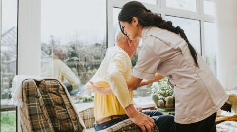 A care worker helps lift an elderly man from an armchair