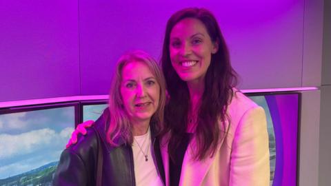 Two women are standing in a purple radio studio. There are tv screens behind them. Both are looking at the camera and smiling. The woman on the right hand side of the photo is wearing a cream-coloured jacket, while the woman on the left hand side of the image is wearing a black leather jacket.