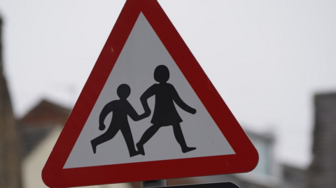 A school sign, crossing patrol warning. The warning sign is in a triangular shape. It has a red border and a white background with two figures portraying a woman and child walking while holding hands.
