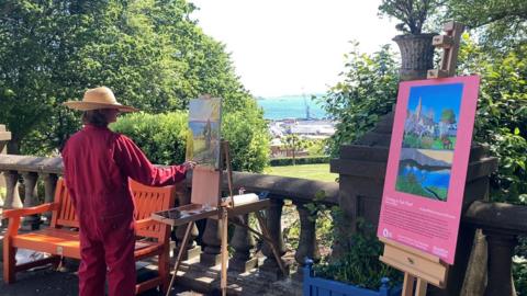 A woman in a red jumpsuit painting in a park.
