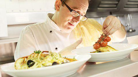 John Quigley in a kitchen. He is dressed in chef's whites, with glasses, and is looking at a plate with food on it in front of him.