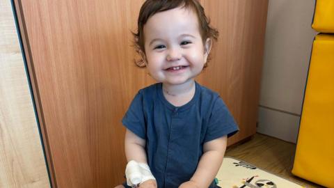 Two-year-old boy with dark hair, wearing a navy playsuit, smiles at the camera.