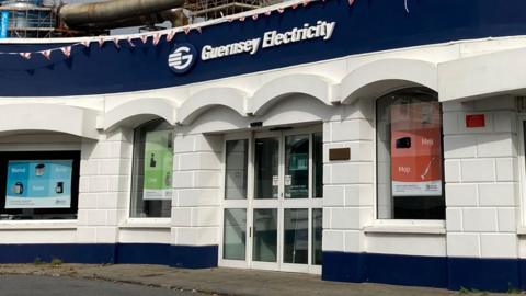 The front building of Guernsey Electricity - the sign above the glass double door, with three windows in vision. The building is painted striped blue and white.