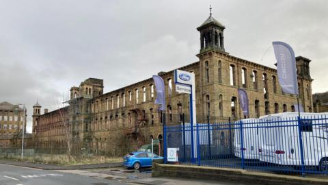 A light-brown coloured shell of a former mill building with scaffolding on two towers at the front of the structure and black charring on another tower's turret evidence of fire damage. 