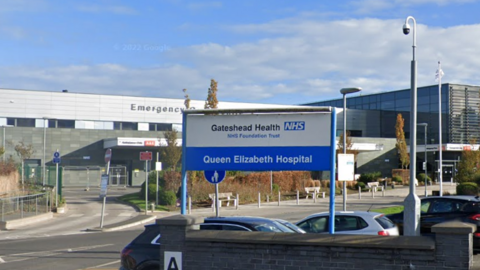 The outside of the Queen Elizabeth Hospital, which is marked with a blue and white sign. Behind is a large, grey building with a car park in the foreground. 