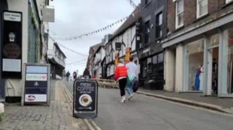 George Street, St Albans with shops on either side, some of which are Tudor styled white with black beams. Two people are walking in the road, one has a red jacket and black trousers and the other is wearing a grey jumper and blue jeans and is carrying a black bag.