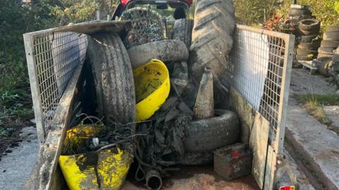 Rubbish, including large tyres, netting, plastics and rubber tubing, piled up in the back of a trailer. The trailer has metal panel on the sides and there is hedges in the background.