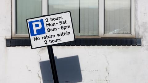 Parking sign with blue square and white P in the middle leans to the left in front of a window with a black sill and a cracked white wall. The shadow of the sign shows underneath it. 