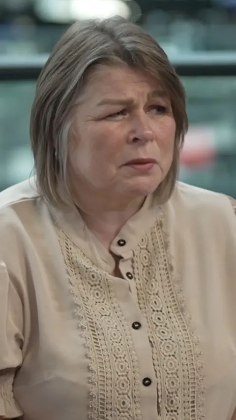 Woman with short brown hair with highlights wearing light beige blouse.