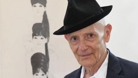 David Remfrey looking at the camera wearing a black trilby hat, white shirt and a dark jacket. Behind him is one of his artworks featuring the 1960s band the Ronettes