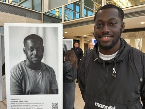 Jason Selormey, a young man with short hair and a beard, smiles as he stands next to his portrait, which is featured in the exhibition. Jason is wearing a black top with a white ribbon pinned to it. He is also wearing a white ribbon in the portrait. The gallery showing the exhibition is in the background and people can be seen looking at the pictures.