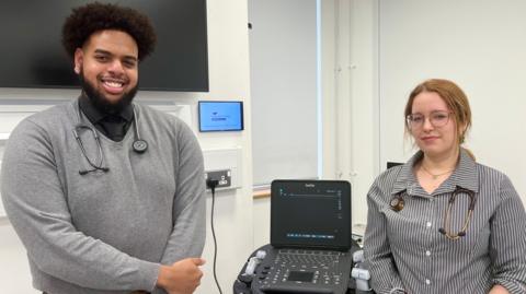 Two medical sudents are standing in a white teaching room, on either side of a piece of electronic medical equipment. The man on the left, who has a beard, is wearing a light grey sweater, with a stethoscope around his neck. The woman on the right has a black and white striped shirt. She also has a stethoscope round her neck and is wearing glasses.