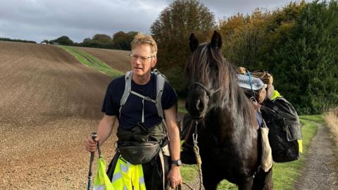 Roger Sewill during a trek in a field next to Scarlet. She is carrying his bags.