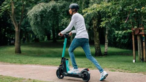 A person with dreadlocks riding a Beryl e-scooter through a park in an overcast day. They are looking away from the camera.
