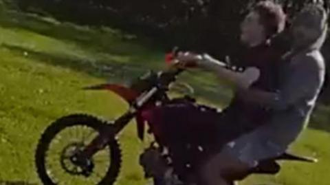 A photograph taken from police bodyworn video showing two young men on a scrambler bike in Stowlawn Green. The one on the front of the bike is holding the handlebars and looking straight forwards. He is wearing  a black T-shirt and black trousers and trainers. The other sitting behind him is wearing shorts and trainers and a grey hoodie and he has turned his head to look at the person taking the image.