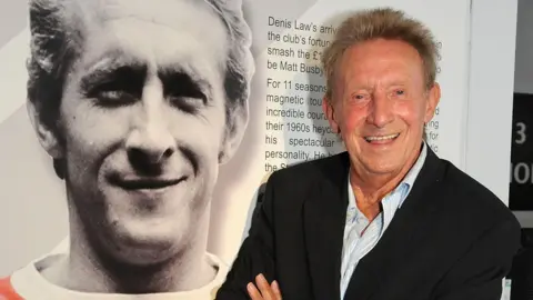 Denis Law standing next to an image of himself as a player during an exhibition at Old Trafford