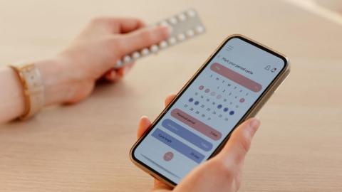 A woman checks her calendar on her mobile phone while holding a packet of contraceptive pills in her other hand