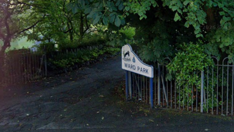 A sign for Ward Park at the side of a road. There is a silver fence and tree covered in green leaves.