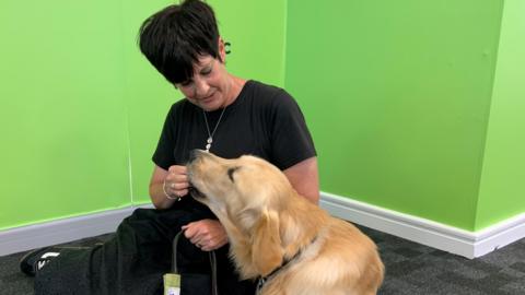 A woman with short black hair is wearing a long black dress. She is sitting on the floor with a golden labrador guide dog and feeding him a treat. There is a bright green wall in the background