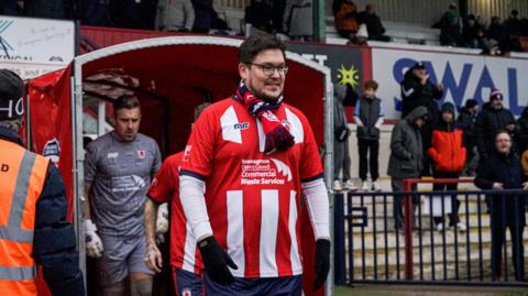 A man in a red and white stripe football shirt walking out from the tunnel onto a football pitch. He is also wearing a red team scarf and black gloves. There is a football player in grey behind him and the crowd can be seen to the right. The man is wearing glasses and is looking to the right of the shot. 