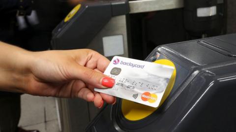 A woman's hand swiping a silver card over a contactless payment point on a barrier.