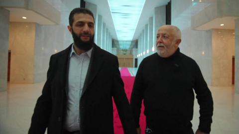 Ahmed al-Sharaa, wearing a black suit, walking with the 鶹Լ's Jeremy Bowen